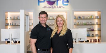 Indoor professional portrait of a man and woman standing together in a clean, modern pharmacy setting. The man on the left has short dark hair and wears a black polo shirt with a small "Pure" logo on the chest, paired with grey trousers. The woman on the right has long, wavy blonde hair and wears a black long-sleeve button-down blouse. They stand in front of a sleek white counter featuring desktop computer monitors. Behind them, glass product display shelves are recessed into white illuminated alcoves. A large backlit sign on the wall above features the company name, "Pure PHARMACY," with a colorful purple mortar and pestle integrated into the lettering.