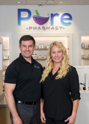 Indoor professional portrait of a man and woman standing together in a clean, modern pharmacy setting. The man on the left has short dark hair and wears a black polo shirt with a small "Pure" logo on the chest, paired with grey trousers. The woman on the right has long, wavy blonde hair and wears a black long-sleeve button-down blouse. They stand in front of a sleek white counter featuring desktop computer monitors. Behind them, glass product display shelves are recessed into white illuminated alcoves. A large backlit sign on the wall above features the company name, "Pure PHARMACY," with a colorful purple mortar and pestle integrated into the lettering.