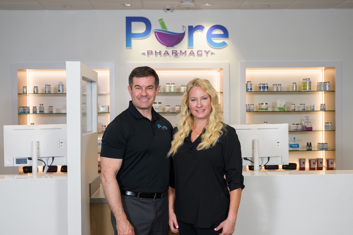 Indoor professional portrait of a man and woman standing together in a clean, modern pharmacy setting. The man on the left has short dark hair and wears a black polo shirt with a small "Pure" logo on the chest, paired with grey trousers. The woman on the right has long, wavy blonde hair and wears a black long-sleeve button-down blouse. They stand in front of a sleek white counter featuring desktop computer monitors. Behind them, glass product display shelves are recessed into white illuminated alcoves. A large backlit sign on the wall above features the company name, "Pure PHARMACY," with a colorful purple mortar and pestle integrated into the lettering.