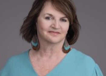 A professional studio headshot of a middle-aged woman with shoulder-length brown hair and a slight smile against a solid grey background. She is wearing a light teal V-neck blouse with small front buttons and large, statement tear-drop-shaped leather earrings featuring a turquoise and brown pattern.