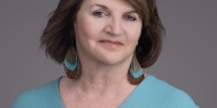A professional studio headshot of a middle-aged woman with shoulder-length brown hair and a slight smile against a solid grey background. She is wearing a light teal V-neck blouse with small front buttons and large, statement tear-drop-shaped leather earrings featuring a turquoise and brown pattern.