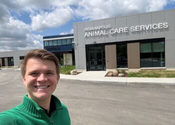 A smiling young man wearing a green quarter-zip sweater takes a selfie outside the modern, newly constructed Indianapolis Animal Care Services building. The grey paneled facility features large glass windows and the agency name in white block letters above the main entrance, with a separate blue "ADOPTIONS" wing visible in the background under a blue sky with scattered clouds.