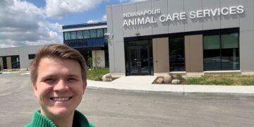 A smiling young man wearing a green quarter-zip sweater takes a selfie outside the modern, newly constructed Indianapolis Animal Care Services building. The grey paneled facility features large glass windows and the agency name in white block letters above the main entrance, with a separate blue "ADOPTIONS" wing visible in the background under a blue sky with scattered clouds.