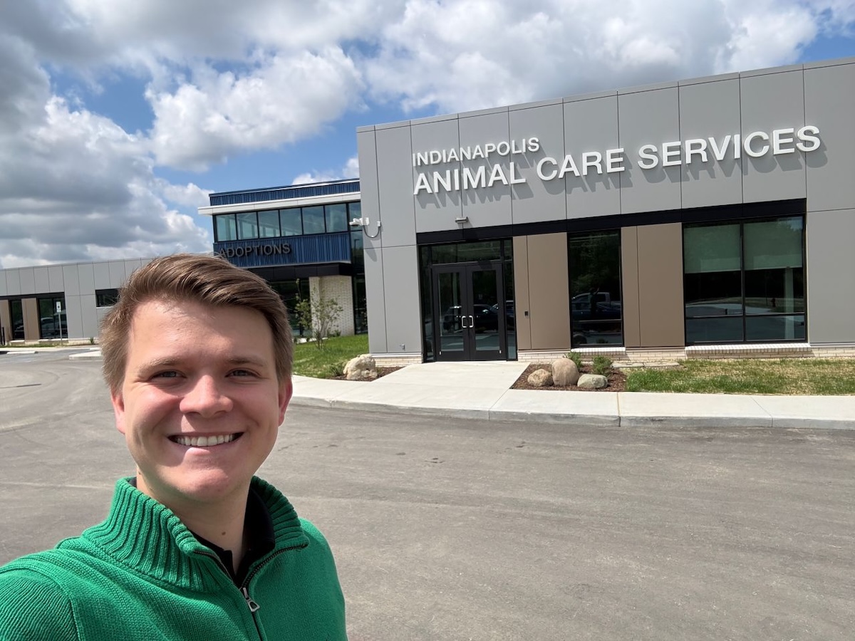 A smiling young man wearing a green quarter-zip sweater takes a selfie outside the modern, newly constructed Indianapolis Animal Care Services building. The grey paneled facility features large glass windows and the agency name in white block letters above the main entrance, with a separate blue "ADOPTIONS" wing visible in the background under a blue sky with scattered clouds.