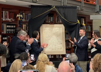 A photograph shows a group of people clapping around a newly unveiled, historical document displayed on an easel. The setting appears to be a library or formal room lined with wooden bookshelves. Two men stand flanking the document under a black drapery frame, while onlookers take photos with their smartphones.