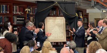 A photograph shows a group of people clapping around a newly unveiled, historical document displayed on an easel. The setting appears to be a library or formal room lined with wooden bookshelves. Two men stand flanking the document under a black drapery frame, while onlookers take photos with their smartphones.
