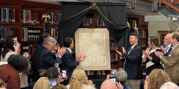 A photograph shows a group of people clapping around a newly unveiled, historical document displayed on an easel. The setting appears to be a library or formal room lined with wooden bookshelves. Two men stand flanking the document under a black drapery frame, while onlookers take photos with their smartphones.