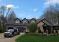A black commercial Ford Super Duty truck parked in the driveway of a large, two-story house with stone and dark gray siding. The home features three roof dormers, an attached garage, and a manicured green lawn with a small tree. A white stepladder stands on the grass near the driveway under a blue sky with light clouds.