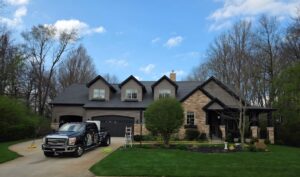 A black commercial Ford Super Duty truck parked in the driveway of a large, two-story house with stone and dark gray siding. The home features three roof dormers, an attached garage, and a manicured green lawn with a small tree. A white stepladder stands on the grass near the driveway under a blue sky with light clouds.