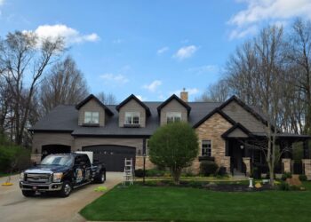 A black commercial Ford Super Duty truck parked in the driveway of a large, two-story house with stone and dark gray siding. The home features three roof dormers, an attached garage, and a manicured green lawn with a small tree. A white stepladder stands on the grass near the driveway under a blue sky with light clouds.