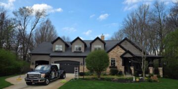 A black commercial Ford Super Duty truck parked in the driveway of a large, two-story house with stone and dark gray siding. The home features three roof dormers, an attached garage, and a manicured green lawn with a small tree. A white stepladder stands on the grass near the driveway under a blue sky with light clouds.