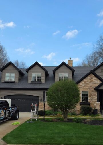 A black commercial Ford Super Duty truck parked in the driveway of a large, two-story house with stone and dark gray siding. The home features three roof dormers, an attached garage, and a manicured green lawn with a small tree. A white stepladder stands on the grass near the driveway under a blue sky with light clouds.
