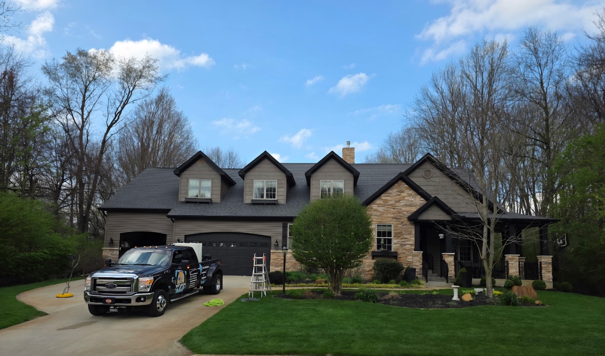 A black commercial Ford Super Duty truck parked in the driveway of a large, two-story house with stone and dark gray siding. The home features three roof dormers, an attached garage, and a manicured green lawn with a small tree. A white stepladder stands on the grass near the driveway under a blue sky with light clouds.