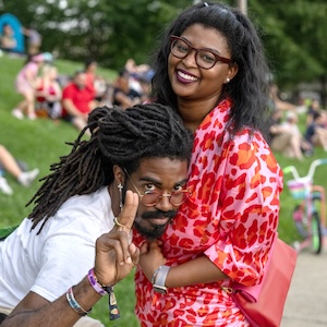 A candid, outdoor portrait shows a smiling Black woman with dark hair and glasses wearing a bright pink and red patterned outfit, while a Black man with long dreadlocks, round glasses, and a mustache leans forward in front of her. He holds up two fingers in a peace sign or gesture toward the camera. They are outdoors at a park, with a grassy hill and blurred onlookers sitting in the background.