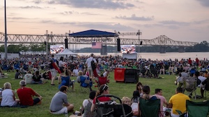 A wide, eye-level outdoor photograph captures a large crowd of people sitting on lawn chairs and blankets on a grassy field, watching a live event on an outdoor stage. The stage is positioned near the edge of a wide river, with a long, continuous steel truss bridge stretching across the background water under a hazy, sunset sky. Large digital screens flank the stage, and several event tents are scattered throughout the venue.