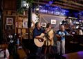 A live music trio performs inside a rustic bar or brewery with wood-paneled walls. On the left, a man sings into a microphone while playing an acoustic guitar. In the center, a woman in a yellow dress plays a smaller acoustic instrument. On the right, a woman in a tie-dye shirt sings into a microphone while playing a mandolin. The background features a bar area, a bar stool, hanging green plants, and a sign on the wall that reads "Muncie."