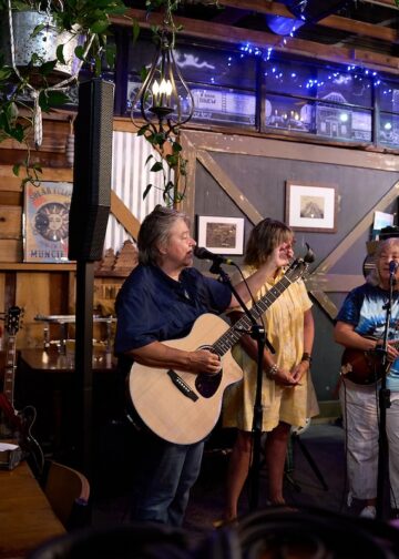 A live music trio performs inside a rustic bar or brewery with wood-paneled walls. On the left, a man sings into a microphone while playing an acoustic guitar. In the center, a woman in a yellow dress plays a smaller acoustic instrument. On the right, a woman in a tie-dye shirt sings into a microphone while playing a mandolin. The background features a bar area, a bar stool, hanging green plants, and a sign on the wall that reads "Muncie."