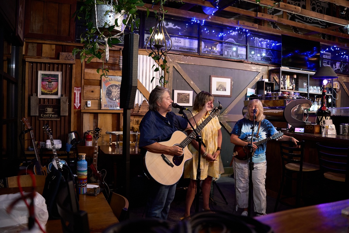 A live music trio performs inside a rustic bar or brewery with wood-paneled walls. On the left, a man sings into a microphone while playing an acoustic guitar. In the center, a woman in a yellow dress plays a smaller acoustic instrument. On the right, a woman in a tie-dye shirt sings into a microphone while playing a mandolin. The background features a bar area, a bar stool, hanging green plants, and a sign on the wall that reads "Muncie."