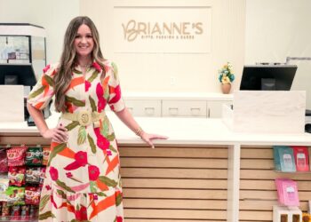 A full-length indoor portrait of a smiling woman standing inside Brianne's Boutique. She has long, wavy light brown hair and is wearing a cream-colored Voy Short Bubble Sleeve Zip Front Floral Midi Dress with a bright red, green, and orange tropical pattern. The dress is cinched at the waist with a wide, woven straw belt featuring a round buckle, and she pairs it with white slip-on slide sandals. She leans her left hand against a long, white checkout counter with light wood slats along the front. In the background, the storefront wall features a large sign that reads "BRIANNE'S GIFTS, FASHION & GARDEN" in gold capital lettering. Gift shop items, including snacks on the left and packaged merchandise on the right, are displayed on retail shelves.
