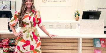 A full-length indoor portrait of a smiling woman standing inside Brianne's Boutique. She has long, wavy light brown hair and is wearing a cream-colored Voy Short Bubble Sleeve Zip Front Floral Midi Dress with a bright red, green, and orange tropical pattern. The dress is cinched at the waist with a wide, woven straw belt featuring a round buckle, and she pairs it with white slip-on slide sandals. She leans her left hand against a long, white checkout counter with light wood slats along the front. In the background, the storefront wall features a large sign that reads "BRIANNE'S GIFTS, FASHION & GARDEN" in gold capital lettering. Gift shop items, including snacks on the left and packaged merchandise on the right, are displayed on retail shelves.