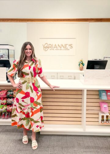 A full-length indoor portrait of a smiling woman standing inside Brianne's Boutique. She has long, wavy light brown hair and is wearing a cream-colored Voy Short Bubble Sleeve Zip Front Floral Midi Dress with a bright red, green, and orange tropical pattern. The dress is cinched at the waist with a wide, woven straw belt featuring a round buckle, and she pairs it with white slip-on slide sandals. She leans her left hand against a long, white checkout counter with light wood slats along the front. In the background, the storefront wall features a large sign that reads "BRIANNE'S GIFTS, FASHION & GARDEN" in gold capital lettering. Gift shop items, including snacks on the left and packaged merchandise on the right, are displayed on retail shelves.
