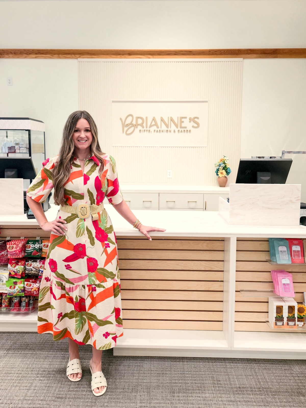 A full-length indoor portrait of a smiling woman standing inside Brianne's Boutique. She has long, wavy light brown hair and is wearing a cream-colored Voy Short Bubble Sleeve Zip Front Floral Midi Dress with a bright red, green, and orange tropical pattern. The dress is cinched at the waist with a wide, woven straw belt featuring a round buckle, and she pairs it with white slip-on slide sandals. She leans her left hand against a long, white checkout counter with light wood slats along the front. In the background, the storefront wall features a large sign that reads "BRIANNE'S GIFTS, FASHION & GARDEN" in gold capital lettering. Gift shop items, including snacks on the left and packaged merchandise on the right, are displayed on retail shelves.
