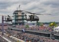 A wide, high-angle shot capturing two military Black Hawk helicopters flying low over the front stretch of the Indianapolis Motor Speedway. In the background, the iconic multi-story Pagoda control tower stands prominently with the "TAG Heuer" clock visible. Massive, packed grandstands filled with thousands of spectators line the track under a cloudy sky. A vertical scoreboard banner reads "110TH INDIANAPOLIS 500". On the track below, race cars and safety vehicles are lined up or moving along pit lane during pre-race ceremonies.