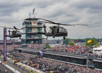 A wide, high-angle shot capturing two military Black Hawk helicopters flying low over the front stretch of the Indianapolis Motor Speedway. In the background, the iconic multi-story Pagoda control tower stands prominently with the "TAG Heuer" clock visible. Massive, packed grandstands filled with thousands of spectators line the track under a cloudy sky. A vertical scoreboard banner reads "110TH INDIANAPOLIS 500". On the track below, race cars and safety vehicles are lined up or moving along pit lane during pre-race ceremonies.