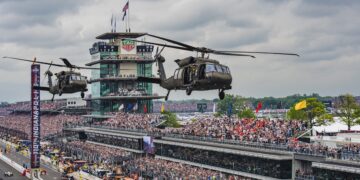 A wide, high-angle shot capturing two military Black Hawk helicopters flying low over the front stretch of the Indianapolis Motor Speedway. In the background, the iconic multi-story Pagoda control tower stands prominently with the "TAG Heuer" clock visible. Massive, packed grandstands filled with thousands of spectators line the track under a cloudy sky. A vertical scoreboard banner reads "110TH INDIANAPOLIS 500". On the track below, race cars and safety vehicles are lined up or moving along pit lane during pre-race ceremonies.