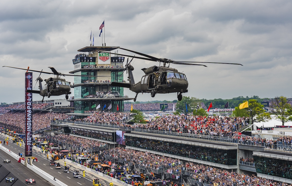 A wide, high-angle shot capturing two military Black Hawk helicopters flying low over the front stretch of the Indianapolis Motor Speedway. In the background, the iconic multi-story Pagoda control tower stands prominently with the "TAG Heuer" clock visible. Massive, packed grandstands filled with thousands of spectators line the track under a cloudy sky. A vertical scoreboard banner reads "110TH INDIANAPOLIS 500". On the track below, race cars and safety vehicles are lined up or moving along pit lane during pre-race ceremonies.