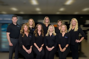 Professional group portrait of eight Pure Pharmacy team members posing together indoors. On the far left, a man stands in a black polo shirt and grey trousers. The remaining seven women stand and sit closely together, all dressed in matching black button-down blouses or polo shirts with a small logo on the left chest. The background is softly blurred, showing a modern office or pharmacy interior with a vending machine or display unit partially visible on the left.