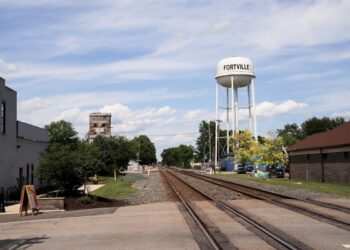 A daytime landscape view in Indiana shows two parallel railroad tracks cutting through Fortville's small downtown area. A large, white metal water tower with "FORTVILLE" painted in black block letters stands tall on the right side of the tracks, next to a brick building. In the distance to the left, an old, weathered concrete grain elevator rises behind trees, under a bright blue sky filled with scattered white clouds.