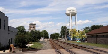 A daytime landscape view in Indiana shows two parallel railroad tracks cutting through Fortville's small downtown area. A large, white metal water tower with "FORTVILLE" painted in black block letters stands tall on the right side of the tracks, next to a brick building. In the distance to the left, an old, weathered concrete grain elevator rises behind trees, under a bright blue sky filled with scattered white clouds.