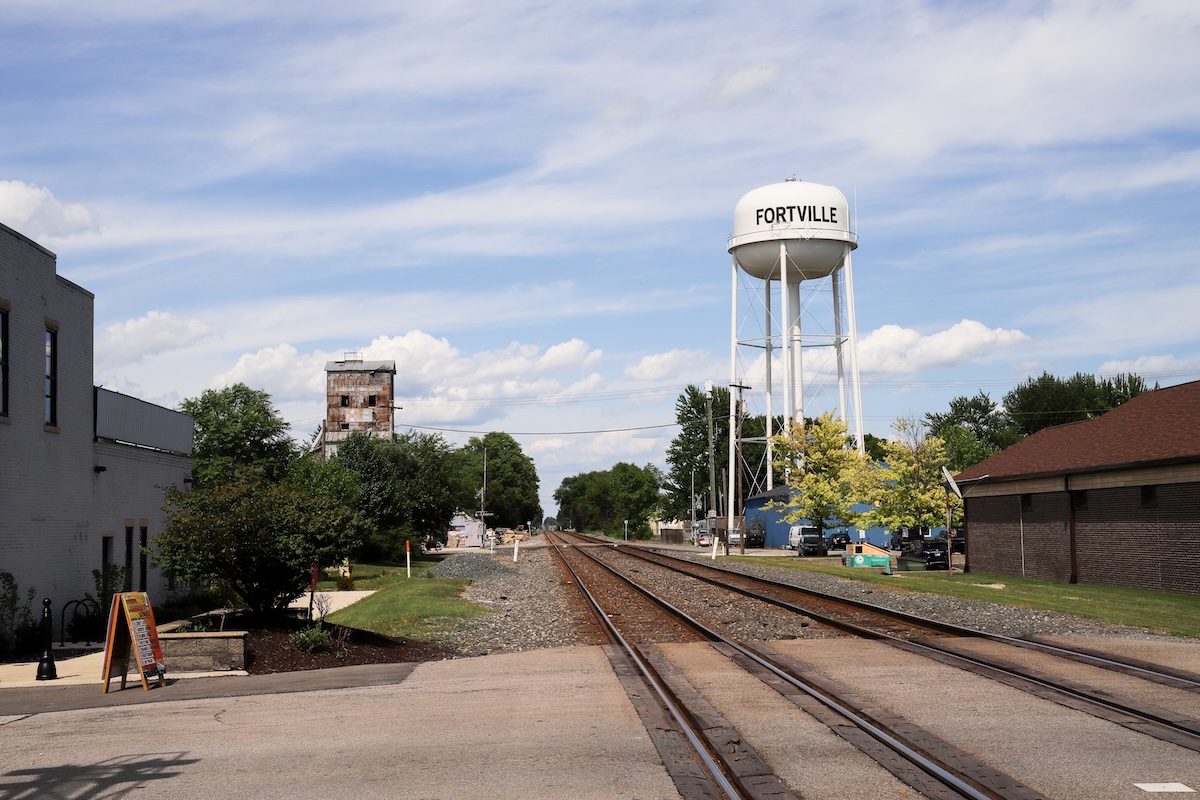 A daytime landscape view in Indiana shows two parallel railroad tracks cutting through Fortville's small downtown area. A large, white metal water tower with "FORTVILLE" painted in black block letters stands tall on the right side of the tracks, next to a brick building. In the distance to the left, an old, weathered concrete grain elevator rises behind trees, under a bright blue sky filled with scattered white clouds.