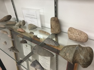 An angled, close-up view of a museum display case filled with ancient stone artifacts. Multiple glass shelves hold various archaeological objects, including primitive stone axe heads, sharpened arrowheads, and carved stone tools. Small informational placards and labels are placed alongside the artifacts, all mounted against a white wall with metal shelving brackets.