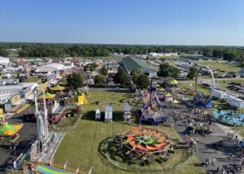 An elevated, high-angle aerial view captures a bustling outdoor county fairground on a sunny day. Various colorful amusement park rides, including a starburst-shaped spinner, a large swinging pendulum ride, and a circular loop ride, are spread across grassy fields and paved walkways. Numerous white event tents, exhibit buildings, and food stalls are clustered on the left, while crowds of people walk along the paths and a massive parking lot filled with cars sits in the background beneath a clear blue sky.