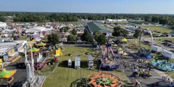 An elevated, high-angle aerial view captures a bustling outdoor county fairground on a sunny day. Various colorful amusement park rides, including a starburst-shaped spinner, a large swinging pendulum ride, and a circular loop ride, are spread across grassy fields and paved walkways. Numerous white event tents, exhibit buildings, and food stalls are clustered on the left, while crowds of people walk along the paths and a massive parking lot filled with cars sits in the background beneath a clear blue sky.