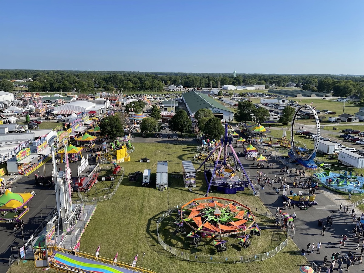 An elevated, high-angle aerial view captures a bustling outdoor county fairground on a sunny day. Various colorful amusement park rides, including a starburst-shaped spinner, a large swinging pendulum ride, and a circular loop ride, are spread across grassy fields and paved walkways. Numerous white event tents, exhibit buildings, and food stalls are clustered on the left, while crowds of people walk along the paths and a massive parking lot filled with cars sits in the background beneath a clear blue sky.