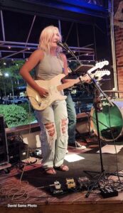 A blonde female musician stands on a small elevated indoor/outdoor stage, singing into a microphone and playing a cream-colored electric guitar. She wears a gray halter-style crop top, heavily distressed light-blue denim jeans with large knee rips, and sandals. Guitar effect pedals sit on the wooden floor at her feet, and a partial view of a green drum kit and another band member is visible behind her, with an open patio window showing cars and bushes outside.