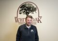 A smiling middle-aged man with a goatee and short brown hair stands indoors in front of a white wall. The wall behind him features a large maroon logo that reads "WHITE OAK CONSTRUCTION" with a silhouette illustration of a large oak tree enclosed in an arch. He wears a black zip-up pullover jacket featuring a matching small White Oak Construction logo on the left chest.