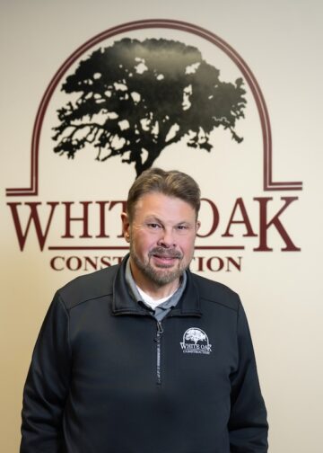 A smiling middle-aged man with a goatee and short brown hair stands indoors in front of a white wall. The wall behind him features a large maroon logo that reads "WHITE OAK CONSTRUCTION" with a silhouette illustration of a large oak tree enclosed in an arch. He wears a black zip-up pullover jacket featuring a matching small White Oak Construction logo on the left chest.