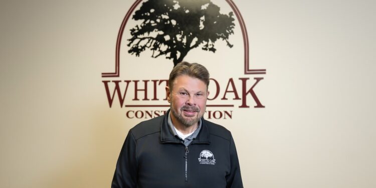 A smiling middle-aged man with a goatee and short brown hair stands indoors in front of a white wall. The wall behind him features a large maroon logo that reads "WHITE OAK CONSTRUCTION" with a silhouette illustration of a large oak tree enclosed in an arch. He wears a black zip-up pullover jacket featuring a matching small White Oak Construction logo on the left chest.