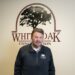 A smiling middle-aged man with a goatee and short brown hair stands indoors in front of a white wall. The wall behind him features a large maroon logo that reads "WHITE OAK CONSTRUCTION" with a silhouette illustration of a large oak tree enclosed in an arch. He wears a black zip-up pullover jacket featuring a matching small White Oak Construction logo on the left chest.