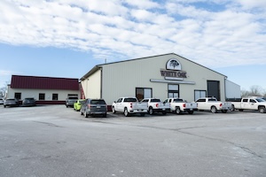 The large white commercial headquarters building of White Oak Construction. A large, prominent oak tree logo sign is mounted above the main entrance. A wide asphalt parking lot in front of the warehouse features a fleet of white commercial pickup trucks, an SUV, and other vehicles under a bright sky with scattered clouds.