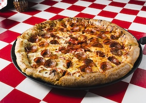 A freshly baked, large pepperoni and cheese pizza sits on a dark metal serving tray over a classic red-and-white checkered tablecloth. The pizza features a thick, golden-brown crust, a melted layer of bubbly mozzarella cheese, and scattered slices of crispy, curled pepperoni.