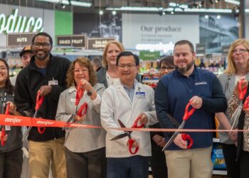 A diverse group of corporate leaders, local officials, and staff stand together inside a grocery store produce department for an official grand opening ribbon-cutting ceremony. They hold large red-handled scissors, cutting a wide red ribbon featuring the orange "Meijer" logo. A man in the center wears a white pharmacy lab coat with a Meijer nametag, while another man to his right wears a blue pullover embroidered with "Brownsburg." In the background, green grocery signs reading "Produce," "Nuts," "Dried Fruit," and "Our Produce" are visible above the displays.