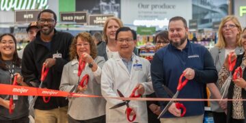 A diverse group of corporate leaders, local officials, and staff stand together inside a grocery store produce department for an official grand opening ribbon-cutting ceremony. They hold large red-handled scissors, cutting a wide red ribbon featuring the orange "Meijer" logo. A man in the center wears a white pharmacy lab coat with a Meijer nametag, while another man to his right wears a blue pullover embroidered with "Brownsburg." In the background, green grocery signs reading "Produce," "Nuts," "Dried Fruit," and "Our Produce" are visible above the displays.