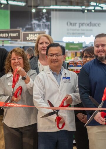A diverse group of corporate leaders, local officials, and staff stand together inside a grocery store produce department for an official grand opening ribbon-cutting ceremony. They hold large red-handled scissors, cutting a wide red ribbon featuring the orange "Meijer" logo. A man in the center wears a white pharmacy lab coat with a Meijer nametag, while another man to his right wears a blue pullover embroidered with "Brownsburg." In the background, green grocery signs reading "Produce," "Nuts," "Dried Fruit," and "Our Produce" are visible above the displays.