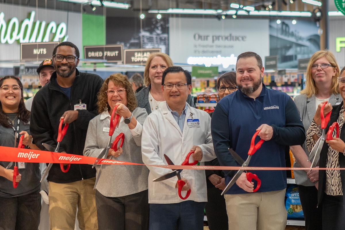 A diverse group of corporate leaders, local officials, and staff stand together inside a grocery store produce department for an official grand opening ribbon-cutting ceremony. They hold large red-handled scissors, cutting a wide red ribbon featuring the orange "Meijer" logo. A man in the center wears a white pharmacy lab coat with a Meijer nametag, while another man to his right wears a blue pullover embroidered with "Brownsburg." In the background, green grocery signs reading "Produce," "Nuts," "Dried Fruit," and "Our Produce" are visible above the displays.