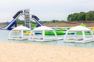 A ground-level, eye-level shot capturing a sandy beach shoreline at SkyLake Adventures in Sheridan, Indiana. In the foreground, three white, inflatable floating cabanas with arched sun canopies sit floating in the calm, light teal water right next to the sand. Each cabana is equipped with comfortable grey lounging pads. In the background across the water, a towering, multi-story gray metal water slide structure stands prominent with long, curving blue and white slides. Vibrant green and yellow inflatable obstacle courses float on the water behind the cabanas under an overcast sky.